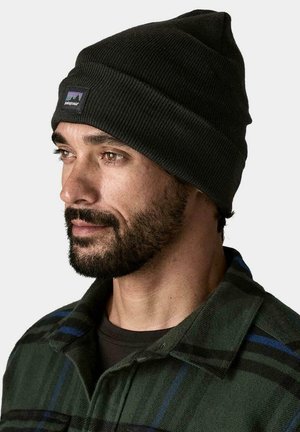 Gorro de punto negro con diseño cónico, que presenta un pequeño parche con un logo colorido. La persona que lo lleva viste una camisa de franela a cuadros en verde y azul.
