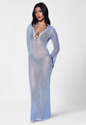 Woman wearing a long, light blue, mesh dress with long sleeves over a bikini, standing with hands behind her back against a plain background.