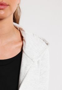 Tejido texturizado de color gris claro con un cuello de chal, superpuesto sobre una blusa negra. Primer plano del área del escote y el hombro.