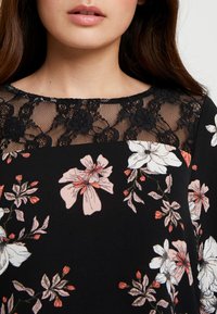 Robe noire à fleurs avec un col en dentelle. Présente de grandes fleurs roses et blanches avec des feuilles vertes, un overlay en dentelle texturée et un tissu doux.