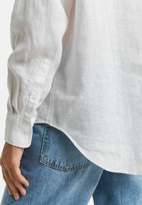 Witte linnen shirt met knoopmanchetten, licht doorzichtig textuur en gekromde zoom. Gepaarde met blauwe spijkerbroek, met zichtbare zakdetails.