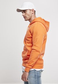 Sudadera con capucha naranja y bolsillo canguro, con cordón. Combinada con jeans de un azul claro y una gorra blanca. Textura de tela suave.