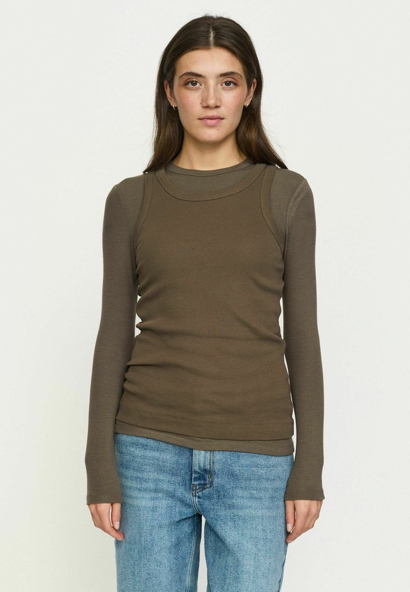 Jeune femme aux longs cheveux foncés porte un haut marron à manches longues superposé et un jean bleu, debout devant un fond clair uni.