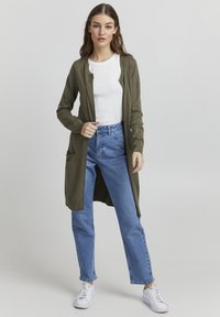 Olive green long cardigan in a textured knit with an open front and two side pockets, paired with light blue straight-leg jeans and white sneakers.