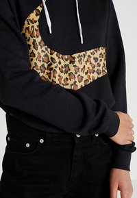 Svart sweatshirt med leopardtryck på swoosh-loggan, vita snoddar och svarta jeans, personens hand vilar på motsatt arm.