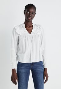 White cotton blouse with ruffled collar and sleeve detailing, featuring pleats and subtle textured accents. Paired with denim jeans.