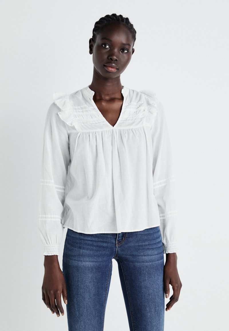 Blusa in cotone bianca con colletto arricciato e dettagli alle maniche, caratterizzata da pieghe e delicati accenti testurizzati. Abbinata a jeans in denim.