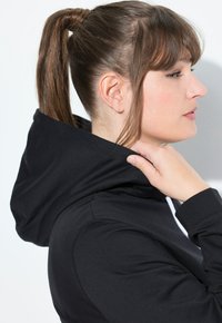 Donna con i capelli castani legati in una coda di cavallo, che indossa un hoodie nero e aggiusta il cappuccio con la mano destra, mostrata di profilo su uno sfondo bianco.