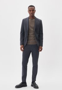 Marineblauer, maßgeschneiderter Anzug mit texturiertem Finish, kombiniert mit einem braunen Strickpullover und schwarzen Loafers. Der Anzug hat einen schlanken Schnitt und ein minimalistisches Design.