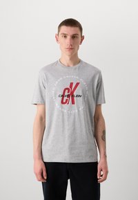 Gray cotton t-shirt featuring a white circular logo with "CALVIN KLEIN" and "CK" in red, short sleeves, crew neckline, relaxed fit.