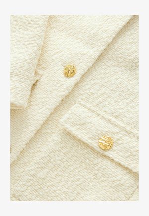 Struktureret cremefarvet stof med et blødt, plysset udseende, der har to runde guldknapper med et distinkt, ribbet design.