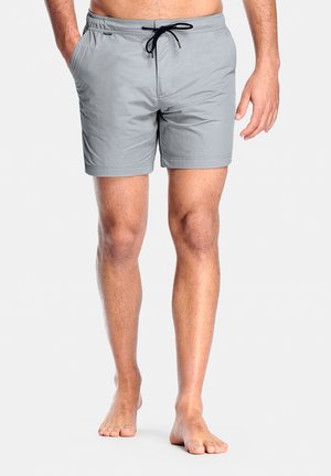 Man draagt lichtgrijze zwembroek met zwarte trekkoord, staat blootsvoets tegen een effen witte achtergrond.