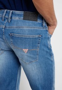 Niebieskie dżinsy z wyblakłym wykończeniem, z skórzaną łatą z logo na pasie i trójkątnym emblematem marki na tylnej kieszeni.