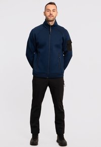 Granatowa polarowa kurtka na zamek z wysokim kołnierzem, czarnymi kieszeniami na zamek i detalami na ramionach; zestawiona z czarnymi spodniami i butami.