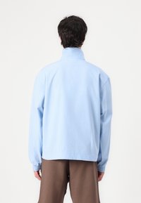 Light blue, oversized jacket with a high collar, textured fabric, and elasticated cuffs. Back view, worn with brown trousers.