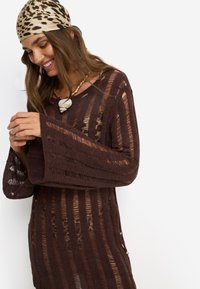 Καφέ πλεκτό τοπ με μακριά, φαρδιά μανίκια και οριζόντιες ρίγες. Διαθέτει υφασμάτινο σχέδιο και μικρή πλευρική λεπτομέρεια με χρυσές αποχρώσεις.