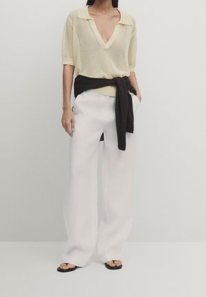 Femme portant un polo en maille jaune clair, un pantalon large blanc, un pull noir noué à la taille, et des tongs noires, debout contre un mur uni.