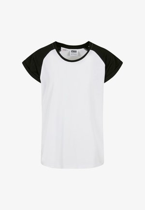 Młoda dziewczynka z długimi blond włosami, ubrana w krótką, białą i czarną koszulę z krótkim rękawem, czarne legginsy i białe sneakersy, stojąca na tle jednolitego tła.