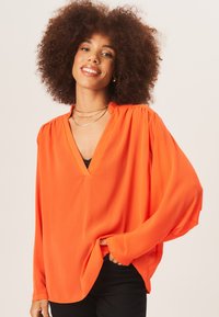 Orange V-neck blouse with long, loose sleeves and gathered shoulders, made of smooth fabric, styled over black pants.
