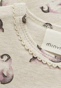 Ljust beige bomullstyg med en mjuk textur, prytt med ett mönster av lila och svarta pingviner; inkluderar en spetsdetalj runt halsringningen.