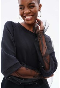 Schwarzer Pullover mit gepufften Ärmeln, der mit durchsichtiger schwarzer Tüll und schwarzen Perlenverzierungen an den Bündchen versehen ist. Bequemer Schnitt mit geripptem Kragen.