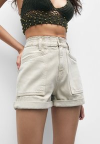 Vaaleanbeige-denim-shortsit, joissa on käärityt lahkeet, etupocketit ja korkea vyötärö. Materiaali näyttää sileältä, hieman karhealta pinnalta.