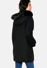 Črn plašč s kapuco, ki ima ovratnik iz umetnega krzna. Narejen je iz teksturirane tkanine in ima sproščen kroj, prikazan s hrbtne strani.