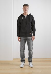 Crna zip-up hoodie s kremastim ušicama, nosi se preko bijele majice, uparen s svijetlo sivim trapericama i bijelim teniscima s crnim prugama.