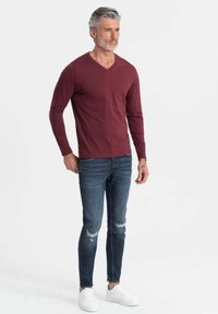 Langarmige, bordeauxrode V-hals shirt gecombineerd met versleten blauwe jeans en witte sneakers. Het shirt heeft een aansluitend ontwerp met een soepele textuur.