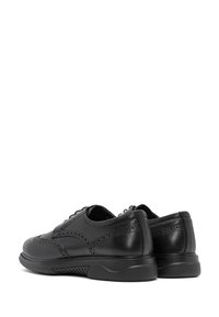 Une paire de chaussures brogue en cuir noir avec des perforations décoratives et des semelles en caoutchouc épaisses, vue de l'arrière et de côté sur fond blanc.