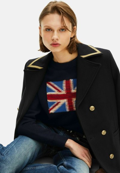 Abrigo negro con cuello acentuado en dorado, combinado con un suéter azul marino que presenta un diseño de la bandera del Reino Unido, y jeans de mezclilla azul. Detalles de botones dorados visibles.