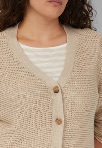 Beige gebreide cardigan met een V-hals en twee houten knopen. De textuur is open gebreid en ligt over een wit en beige gestreept shirt.