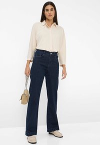 Femme portant un chemisier crème à manches longues, un jean large taille haute bleu foncé, des mocassins beiges, tenant un sac à main beige avec une chaîne.