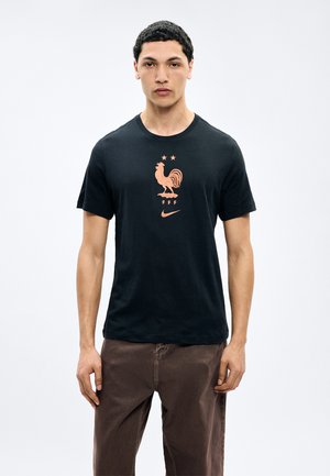 Ung mand iført sort T-shirt med orange hane, to stjerner, "FFF" og Nike-logo, kombineret med brune bukser, stående mod en hvid baggrund.