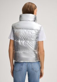 Srebrna kamizelka puffer z wysokim kołnierzem, pikowanym wzorem i błyszczącą teksturą, noszona na białej koszulce, w połączeniu z niebieskimi dżinsami.