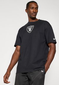 Crna pamučna majica s bijelim logom Raiders na prednjoj strani, kratkim rukavima i ležernim krojem. Uparena s crnim kratkim hlačama s uzorkom.