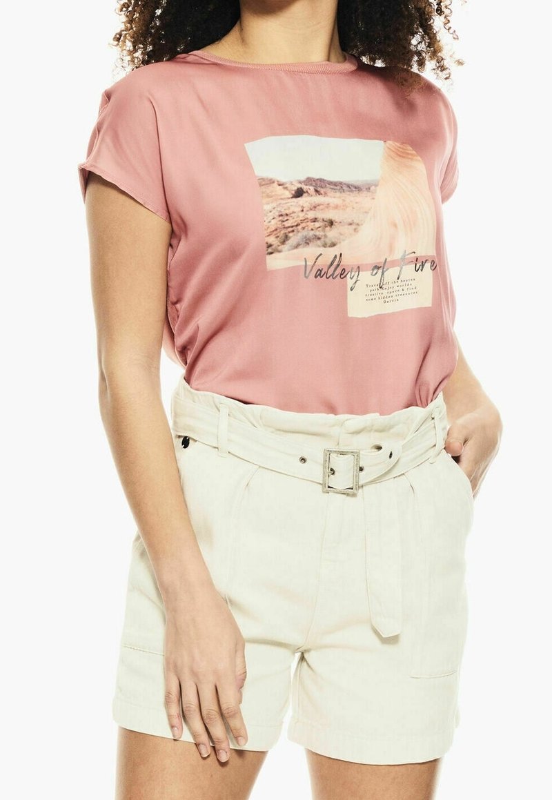 Růžový top s krátkým rukávem s grafickým motivem krajiny a textem "Údolí ohně", spárovaný s bílými kraťasy na opasek, lehký materiál, volný střih.