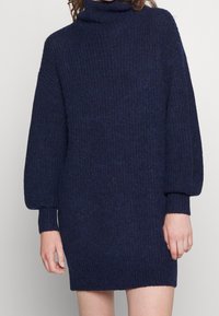 Mörkblå stickad klänning med hög krage, ribsstruktur och ballongärmar, med avslappnad passform och längd till mitten av låret.