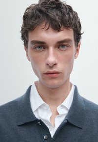 Pletený šedý svetr s límečkem, nošený přes bílé pruhované tričko. Modelka má krátké vlnité vlasy a modré oči.