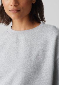 Grauer Pullover mit Rundhalsausschnitt, weicher Textur, mit einem kleinen gestickten Herzen in hellerem Grau auf der Brust.