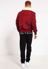 Rød bomberjakke med en faux fur krave, ribstrikkede manchetter og kant. Båret med sorte sweatpants og hvide sneakers, set fra ryggen.