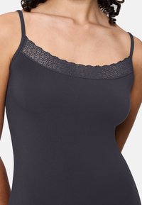 Kobieta ubrana w ciemnoszary top na ramiączkach z koronkowym wykończeniem przy dekolcie, zbliżenie na górną część torsu i ramiona.