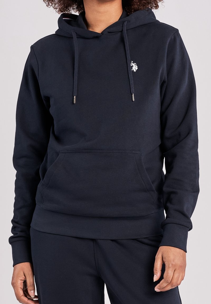 Personne portant un sweat à capuche bleu marine foncé avec une poche avant et un logo blanc brodé sur la poitrine, associé à un pantalon bleu marine assorti.