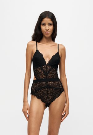 Femme debout pieds nus portant un body en dentelle noire avec fines bretelles sur un fond clair uni.