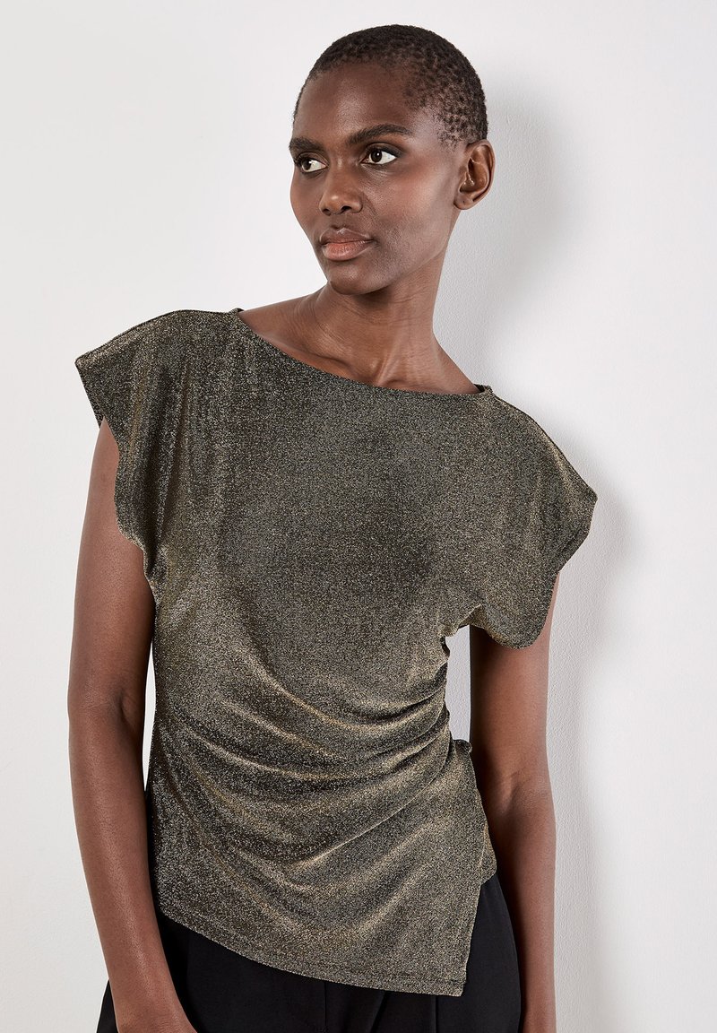 Top metallico oro con maniche fluttuanti corte, scollatura ampia e texture arricciata lungo il lato. Indossato sopra pantaloni neri.