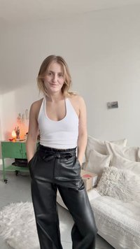 Balts halter top papildināts ar melnām mākslīgās ādas platām biksēm; biksēm ir sasienama josta. Neitrāla iekštelpu vide ar mīkstiem mēbelēm.