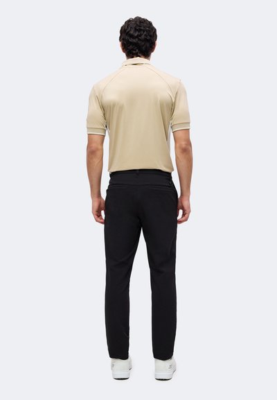Homme debout, dos à la caméra, portant un polo beige à manches courtes, un pantalon noir et des baskets blanches sur un fond uni.