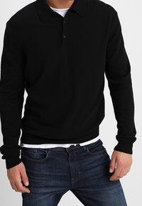 Svart tröja med krage, långa ärmar, ribbade manschetter och nederkant. Bärs med blå jeans. Tyget verkar slätt och lätt.