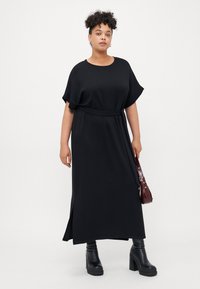 Black, short-sleeved dress with a belted waist and side slit. Styled with black platform boots and a burgundy handbag. Smooth texture.