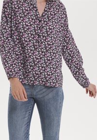 Blouse fleurie en tissu noir avec des fleurs roses et blanches, dotée de manches longues bouffantes et d'un décolleté en V. Associée à un jean bleu.